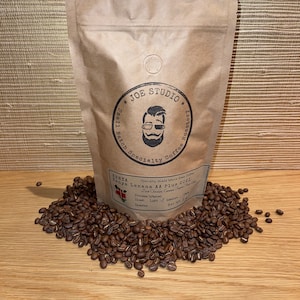 May include: A brown paper bag of Joe Studio Kenya Lenana AA Plus 2021 coffee beans, surrounded by roasted coffee beans. The bag has a logo and text that reads "Small Batch Specialty Coffee Roastery". The bag's label details the coffee's origin and roast.