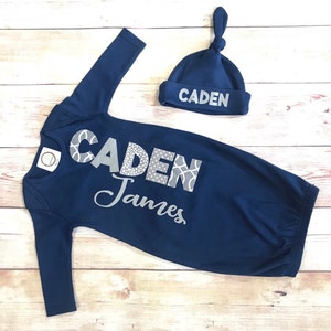 May include: A navy blue baby gown with long sleeves and a matching navy blue hat. The gown has the name "CADEN James" written in silver glitter on the front. The hat has the name "CADEN" written in silver glitter on the front.