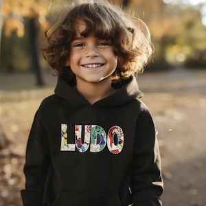 A young boy wearing a black hooded sweatshirt, with his name (LUDO) across the front in a Marvel superhero fabric.