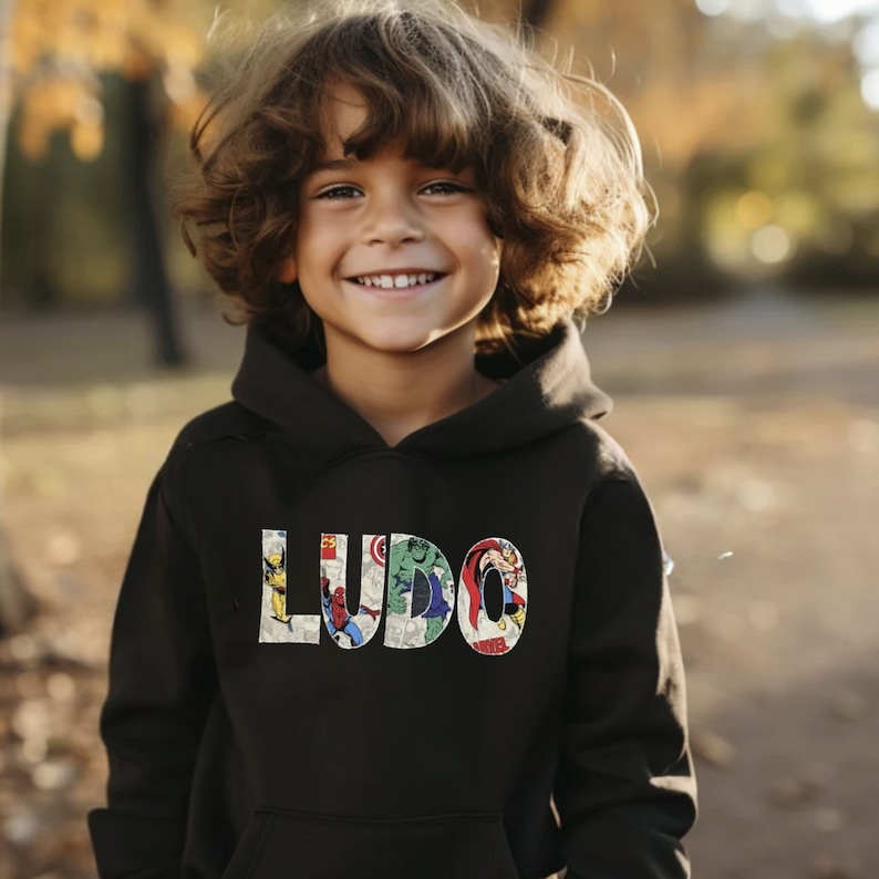 A young boy wearing a black hooded sweatshirt, with his name (LUDO) across the front in a Marvel superhero fabric.