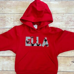 May include: A red hooded sweatshirt featuring the name "ELLA" across the chest. The letters are grey with Minnie Mouse head designs. The sweatshirt has a front pocket and a drawstring hood.