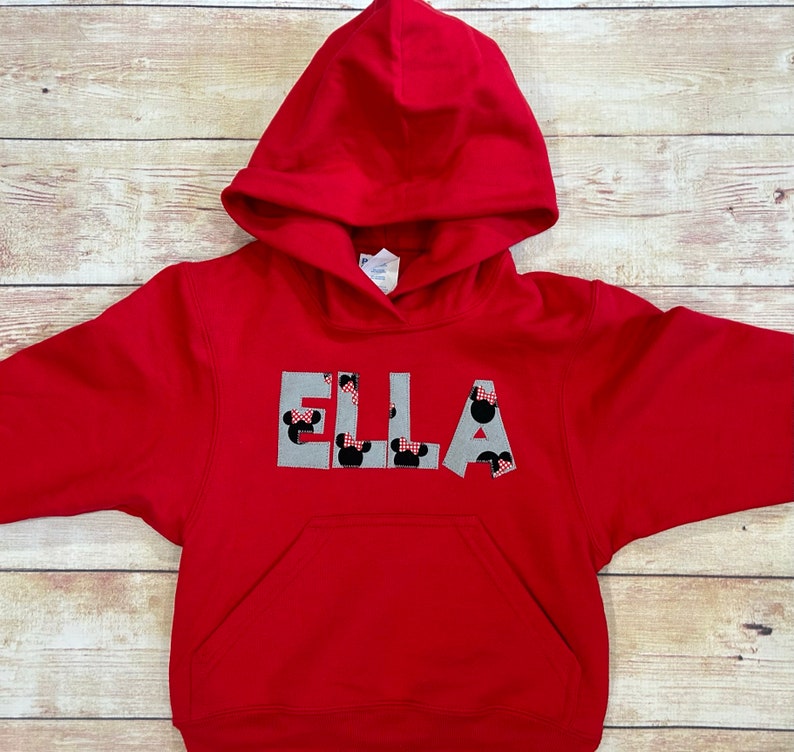 May include: A red hooded sweatshirt featuring the name "ELLA" across the chest. The letters are grey with Minnie Mouse head designs. The sweatshirt has a front pocket and a drawstring hood.