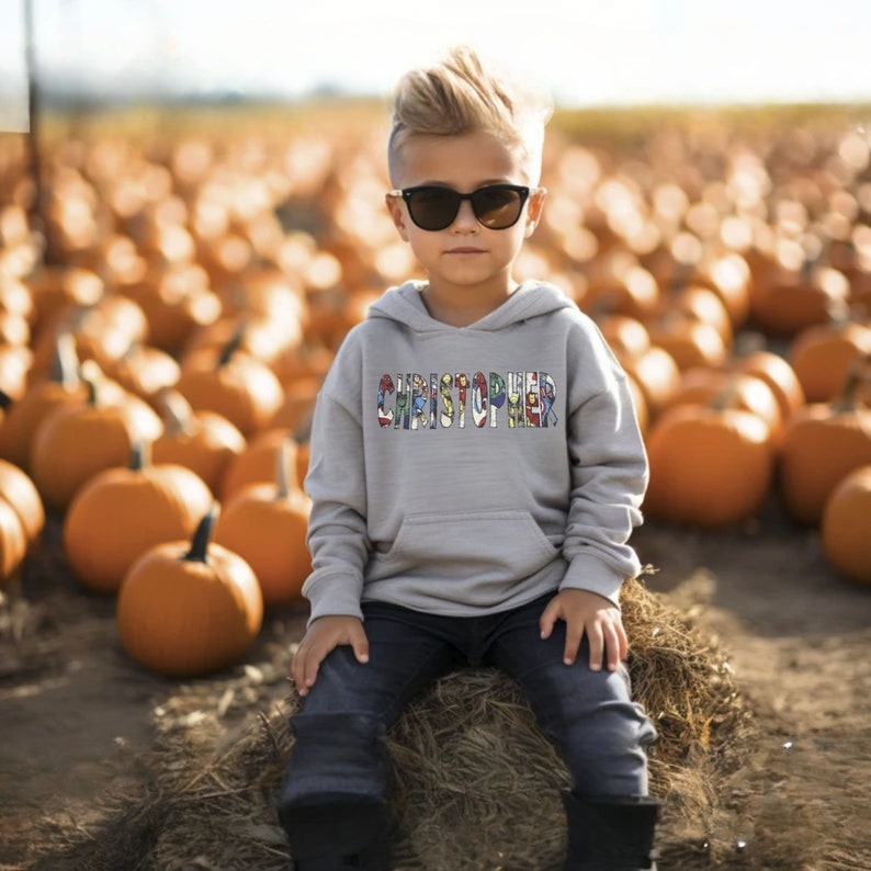 May include: A young boy wearing a gray hoodie with the name "CHRISTOPHER" spelled out in colorful letters. He is wearing blue jeans and sunglasses and sitting on a bale of hay in a field of pumpkins.