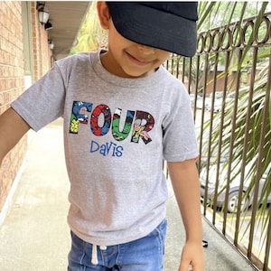 May include: A gray t-shirt with a graphic of the word "FOUR" made up of images of superheroes. The name "Davis" is printed below the graphic.