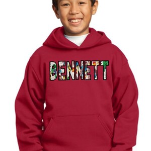 May include: Red hooded sweatshirt with a pocket on the front. The name "BENNETT" is printed on the front in black with a colorful comic book design.