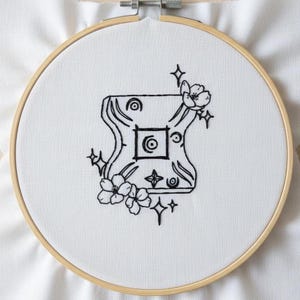 May include: An embroidery hoop with a white fabric featuring a black, stylized design. The design includes a central square with a circle, surrounded by curved lines, flowers, and star-like shapes. The wooden hoop holds the fabric taut.