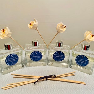 May include: Four glass bottles of natural reed diffuser with decorative flower accents. The bottles are labeled with the text "Crescent Moon" and different scents: Macintosh Apple, Black Currant & Apple, White Pine, and Gingerbread.