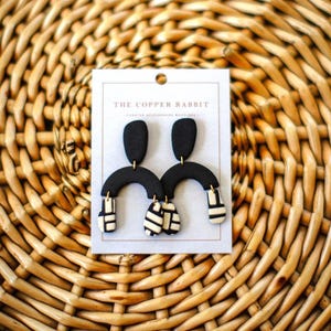 May include: Black and white abstract design earrings. The earrings have a black top, a curved black middle, and a white and black patterned bottom. The earrings are displayed on a white card with the text "THE COPPER RABBIT". The background is a woven basket.