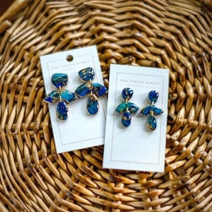 May include: Two pairs of handmade earrings on white cards, displayed on a woven basket. The earrings feature a blue and gold marbled design with a fan-shaped top and a teardrop-shaped bottom. The text "THE COPPER RABBIT" is printed on the cards.