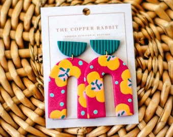 May include: A pair of vibrant, handmade earrings featuring a pink, floral design with yellow and teal accents. The earrings have a unique arch shape and are displayed on a white card with the text "THE COPPER RABBIT".