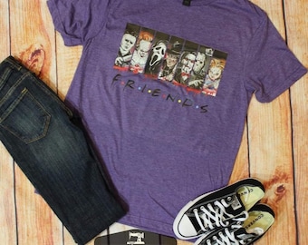 May include: Purple t-shirt with a graphic of horror movie characters in a "Friends" style logo. The text "FRIENDS" is printed below the graphic. The shirt is paired with blue jeans and black sneakers.