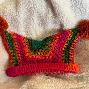 May include: A crocheted beanie featuring a vibrant, multi-colored striped pattern in shades of hot pink, orange, dark green, and brown. The hat has a wide, ribbed brim and two large, round, orange yarn pom-poms attached to the top corners.