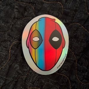 May include: A holographic sticker featuring a stylised illustration of Deadpool's face. The left side of the face is a rainbow gradient, and the right side is red. The sticker has the text "Etsy @thewealdierthewind" at the bottom.
