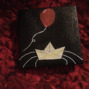 Könnte beinhalten: Ein schwarzes Leinwandbild von einem roten Ballon, der an einem weißen Papierboot befestigt ist, auf dem die Worte "S.S. Georgie" stehen.