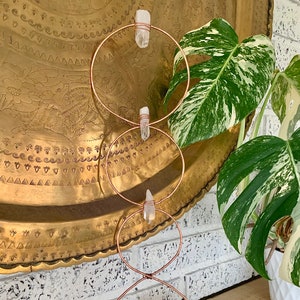 May include: A copper wire stand with three circular hoops, each holding a clear quartz crystal. The stand is placed on a wooden surface with an amethyst crystal cluster in the foreground.