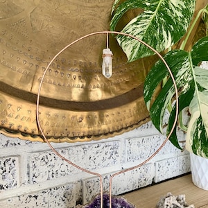 May include: A copper wire crystal holder with a clear quartz crystal hanging from the top. The holder is standing on a wooden surface with a purple amethyst crystal and a white crystal next to it.
