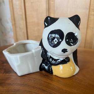 May include: A ceramic planter shaped like a panda bear holding a yellow ball. The planter is white with black markings and has a separate heart-shaped planter attached to the side.