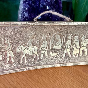 May include: A silver metal wall plaque with a raised design of a procession of people and animals. The plaque has a textured surface and a decorative border.