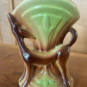 May include: A ceramic vase shaped like a deer with a green and brown glaze. The deer is standing on its hind legs with its head tilted back. The vase has a wide opening at the top and a narrow base.