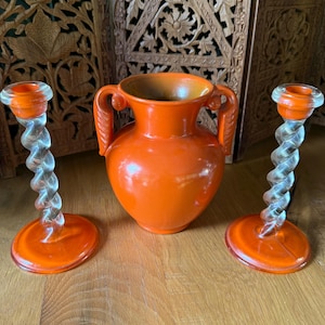 May include: A set consisting of one orange ceramic vase with two side handles and two clear glass candlesticks. Each candlestick features a twisted stem design and a solid orange circular base, matching the color of the central vase.