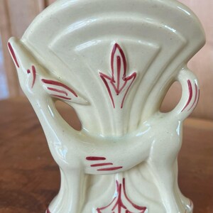 May include: A cream-colored ceramic vase with a stylized deer design and red accents. The vase has a wide, fan-shaped opening and a rounded base.