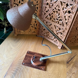 May include: A brown desk lamp with a textured lampshade and a brass-colored adjustable arm. The lamp sits on a square wooden base with a circular accent. The lamp is a vintage style.