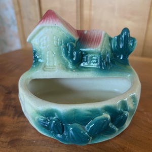 May include: A small ceramic planter shaped like a house with a green and white exterior and a red roof. The planter has a small opening in the front for planting. The planter is decorated with green leaves and a small tree on the side.