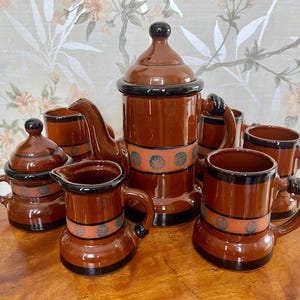 May include: A brown ceramic tea set with a teapot, creamer, sugar bowl, and four mugs. The set features a black trim and a decorative pattern of orange circles with blue accents.