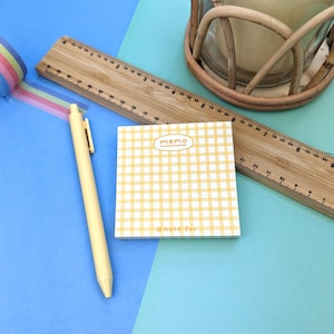 Puede incluir: Un bloc de notas cuadrado con estampado de cuadros vichy amarillos y blancos con el texto memo en la parte superior y note for en la inferior, junto a un bolígrafo retráctil amarillo claro y una regla de madera de 30 cm.