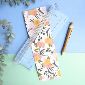 Puede incluir: Una guillotina de papel azul claro con una gu&iacute;a de corte de pl&aacute;stico transparente, colocada sobre una tarjeta rectangular con un patr&oacute;n floral de flores amarillas, melocot&oacute;n y rosas, hojas verdes y peque&ntilde;os p&aacute;jaros azules. Un bol&iacute;grafo de madera a la derecha.