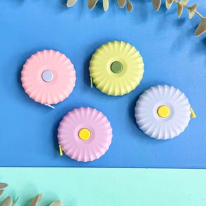 May include: Four circular retractable measuring tapes with a scalloped edge design. The tapes are coloured pastel pink, pale yellow, lavender, and light purple, each featuring a small contrasting centre button in light blue, olive green, or yellow.