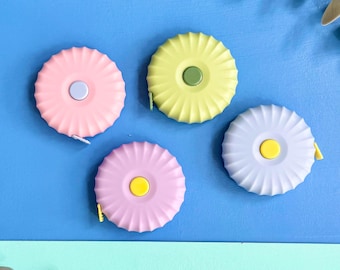 Puede incluir: Cuatro cintas métricas retráctiles circulares con un diseño de borde festoneado. Las cintas son de color rosa pastel, amarillo pálido, lavanda y violeta claro, cada una con un pequeño botón central en contraste azul claro, verde oliva o amarillo.