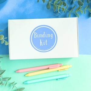Puede incluir: Una caja blanca con un círculo azul que dice "Binding Kit" y el sitio web "cornflowerstone.com". Tres bolígrafos de colores pastel están sobre una superficie verde claro.