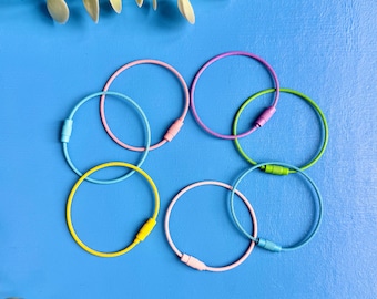 Puede incluir: Un conjunto de siete llaveros circulares de alambre en varios colores, incluyendo rosa, azul claro, amarillo, morado y verde. Cada anillo cuenta con un conector de barril metálico roscado que permite abrir y cerrar el bucle de forma segura.