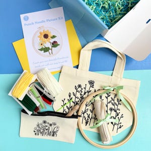 May include: A punch needle picture kit containing a printed instruction card, a beige canvas tote bag with black floral line art, a matching pencil case, a wooden embroidery hoop, a roll of fabric, colourful yarn skeins, and a needle tool.