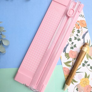 Puede incluir: Una guillotina de papel rosa claro con una gu&iacute;a de corte de pl&aacute;stico transparente, un mecanismo de cuchilla deslizante y una cuadr&iacute;cula blanca impresa en la base. Un bol&iacute;grafo de bamb&uacute; descansa al lado sobre un papel con estampado floral.