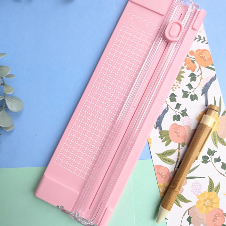 Puede incluir: Una guillotina de papel rosa claro con una gu&iacute;a de corte de pl&aacute;stico transparente, un mecanismo de cuchilla deslizante y una cuadr&iacute;cula blanca impresa en la base. Un bol&iacute;grafo de bamb&uacute; descansa al lado sobre un papel con estampado floral.