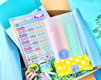 May include: A box filled with stationery supplies, including a teal notebook, stickers, washi tape, and a yellow polka-dot pouch. The stickers have the days of the week printed on them. The box is lined with blue and green tissue paper.