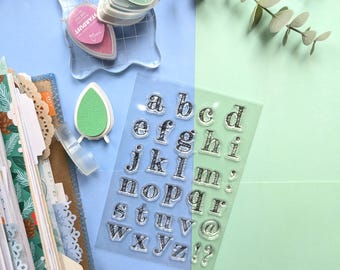 May include: A clear sheet of lowercase alphabet stamps with a plaid pattern, alongside a clear acrylic stamping block, a pink Stardust ink pad, a light green ink pad, and a partially visible planner with patterned pages and decorative paper edges.