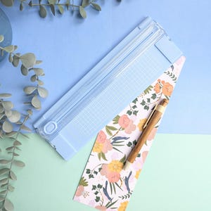 Puede incluir: Una guillotina de papel azul claro con una base cuadriculada, colocada en diagonal junto a una tira rectangular de papel con un estampado floral en tonos rosa, amarillo y verde, y un bol&iacute;grafo de madera con punta met&aacute;lica sobre el papel.