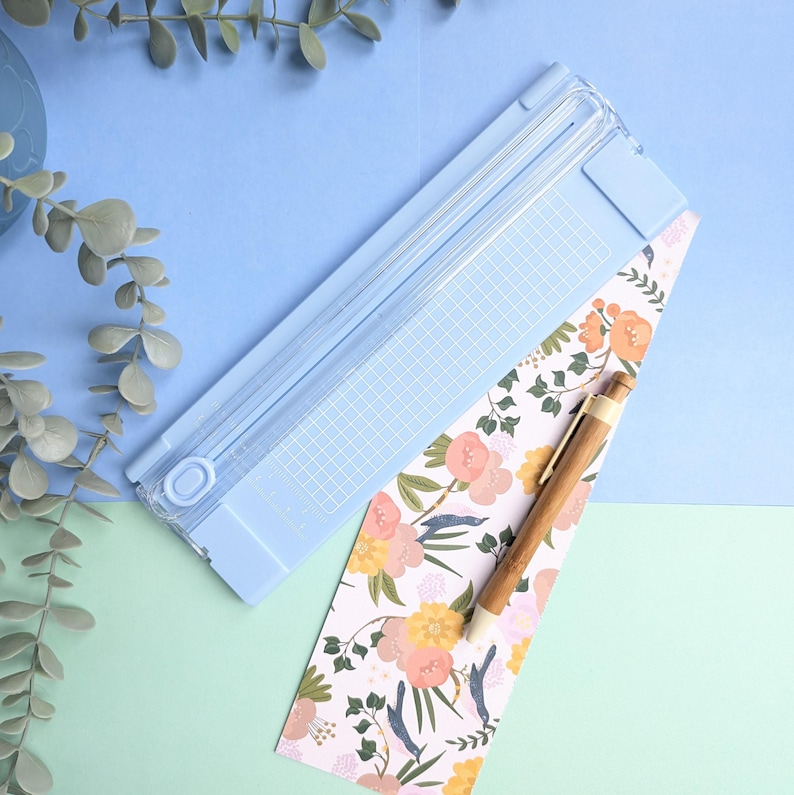 Puede incluir: Una guillotina de papel azul claro con una base cuadriculada, colocada en diagonal junto a una tira rectangular de papel con un estampado floral en tonos rosa, amarillo y verde, y un bol&iacute;grafo de madera con punta met&aacute;lica sobre el papel.