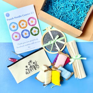May include: A punch needle coaster kit including a printed instruction card, a wooden embroidery hoop with grey felt, a canvas zip pouch with a floral print, various spools of colourful yarn, a punch needle tool, and a roll of fabric.