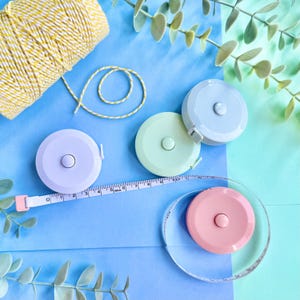 Puede incluir: Una colección de cintas métricas retráctiles de colores pastel, incluyendo lavanda, verde menta y rosa, con cintas métricas blancas extendidas. También hay hilo amarillo y blanco y vegetación sobre un fondo azul y verde.