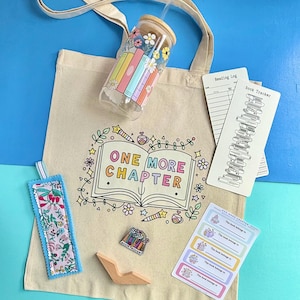 May include: A beige canvas tote bag featuring an open book graphic with the text ONE MORE CHAPTER, a glass tumbler with a bamboo lid, a floral fabric bookmark, a book tracker card, a small enamel pin, and a sheet of book ownership labels.