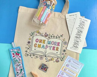 May include: A beige canvas tote bag featuring an open book graphic with the text ONE MORE CHAPTER, a glass tumbler with a bamboo lid, a floral fabric bookmark, a book tracker card, a small enamel pin, and a sheet of book ownership labels.