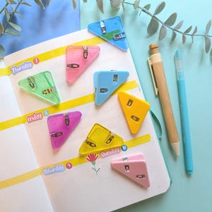May include: A collection of triangular, translucent plastic corner clips in various colors including pink, blue, yellow, and green, placed on an open planner page with yellow washi tape, a wooden mechanical pencil, and a light blue gel pen.