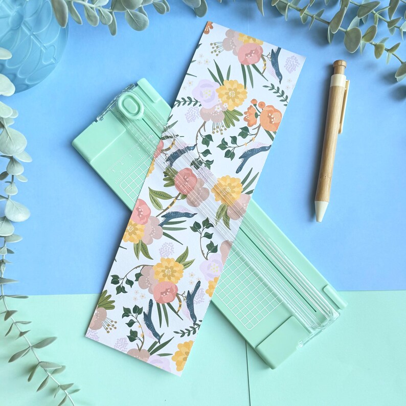 Puede incluir: Una guillotina de papel verde menta con una gu&iacute;a de corte de pl&aacute;stico transparente se sit&uacute;a en diagonal sobre un marcap&aacute;ginas rectangular con un estampado floral de flores amarillas, rosas y lavanda con hojas verdes. Un bol&iacute;grafo de bamb&uacute; a la derecha.