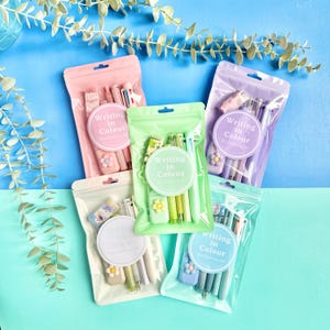 May include: Five stationery sets in resealable pouches, each containing pens and an eraser with a flower accent. The circular labels on the front of each pouch feature the text Writing in Colour. The sets are pink, green, purple, white, and blue.