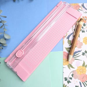 Puede incluir: Una guillotina de papel rosa claro con una gu&iacute;a de corte de pl&aacute;stico transparente, un mecanismo de cuchilla deslizante y una superficie con cuadr&iacute;cula impresa. Un bol&iacute;grafo de bamb&uacute; con punta blanca descansa sobre papel floral.
