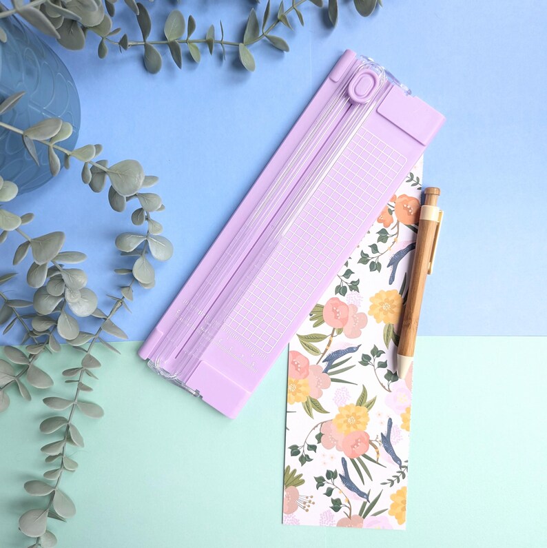 Puede incluir: Una guillotina de papel de color violeta claro con una gu&iacute;a de corte de pl&aacute;stico transparente y una superficie cuadriculada, colocada junto a un marcap&aacute;ginas rectangular con un patr&oacute;n floral de flores rosas y amarillas y hojas verdes, y un bol&iacute;grafo de madera.